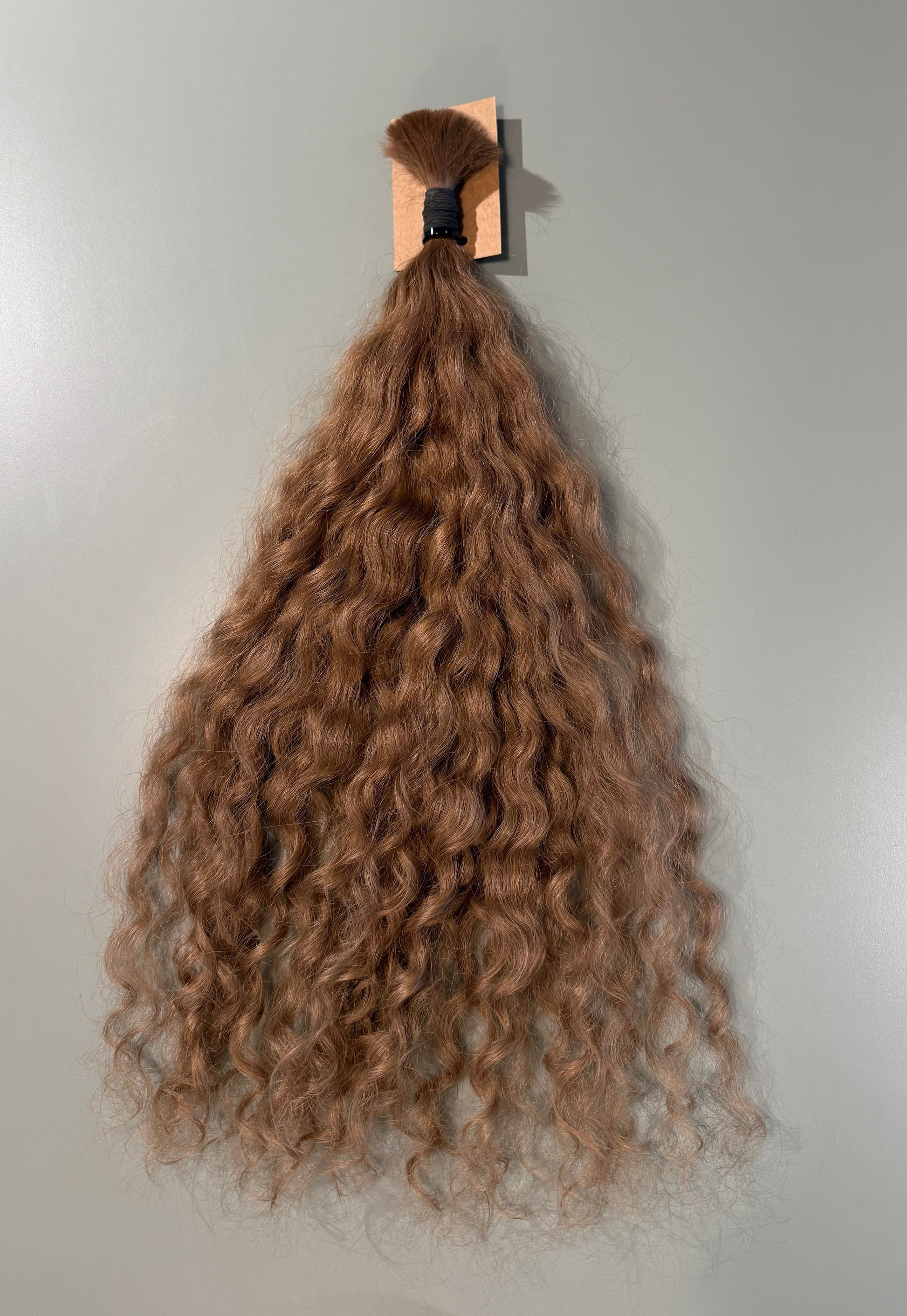 A bundle of long, curly Slavic hair extensions in a light brown shade, hanging from a wooden stand against a plain light gray wall.