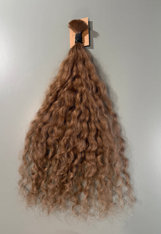 A bundle of long, curly Slavic hair extensions in a light brown shade, hanging from a wooden stand against a plain light gray wall.