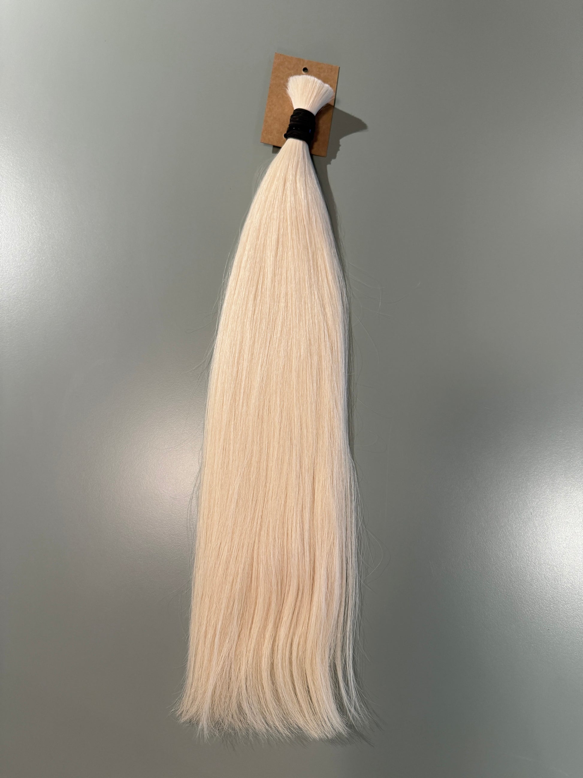 A single bundle of long, straight slavic hair extensions in a very light platinum blonde color, attached to a small brown cardboard header with a black elastic band, shown against a plain gray background.