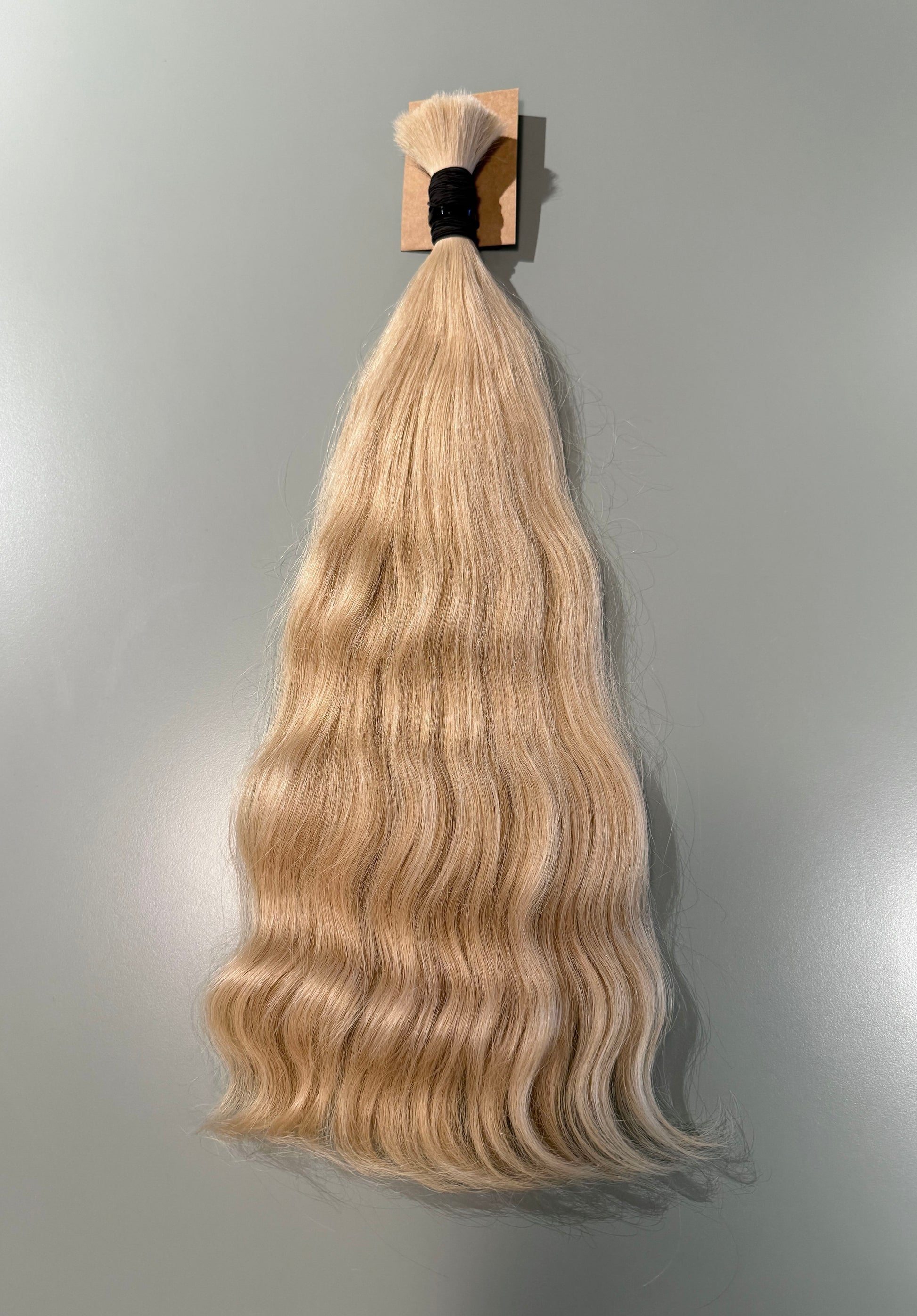 A bundle of long, wavy blonde Slavic hair extensions hanging from a small wooden stand against a plain gray wall.