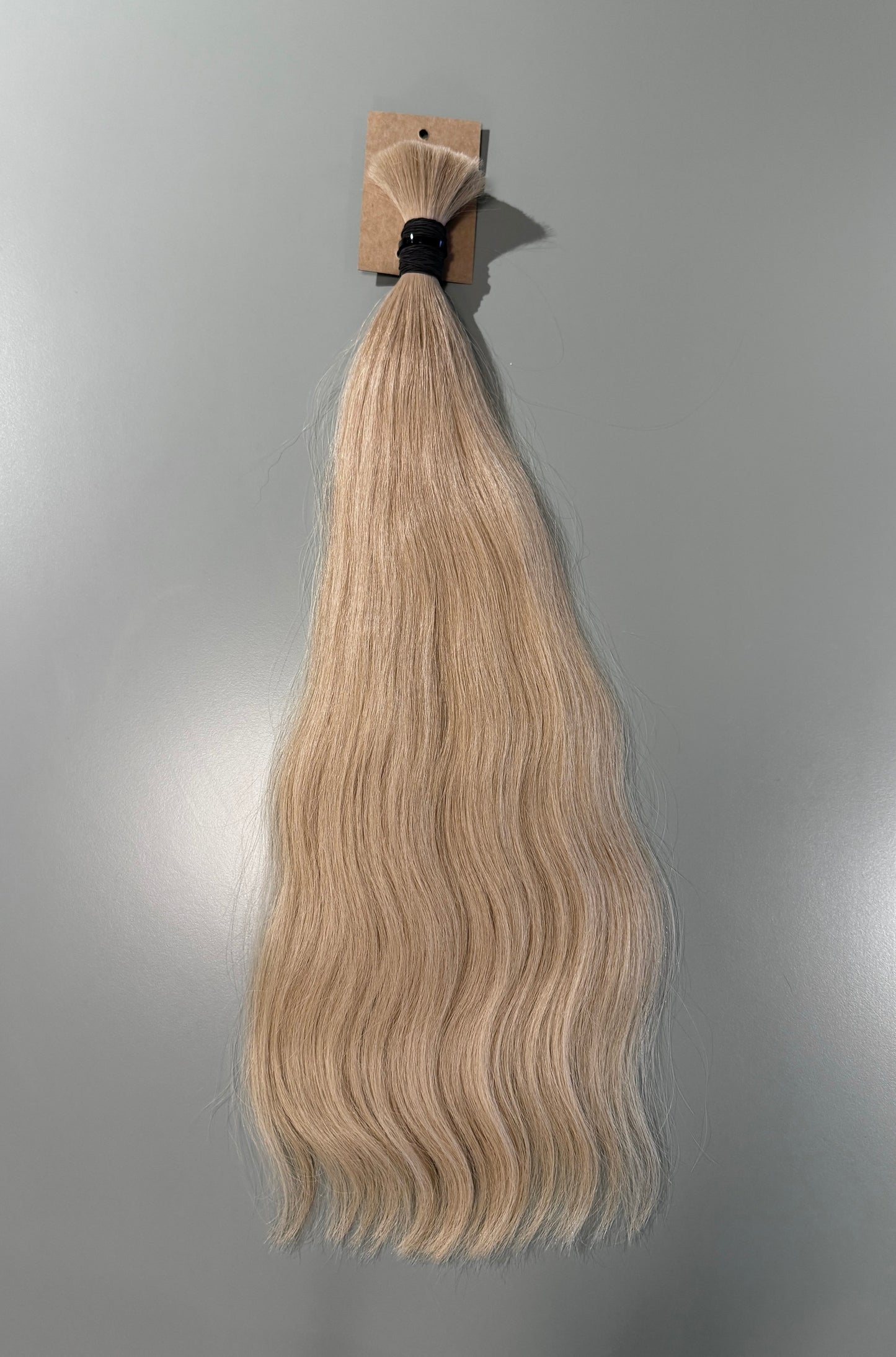 A bundle of wavy blonde Slavic hair extensions attached to a cardboard header card with a black elastic band, shown against a plain gray background.