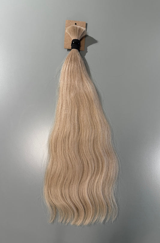A bundle of wavy blonde Slavic hair extensions attached to a cardboard header card with a black elastic band, shown against a plain gray background.