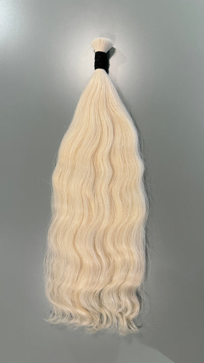 A single bundle of long, wavy slavic hair extensions in a very light platinum blonde color, tied with a black band at the top, set against a plain gray background.