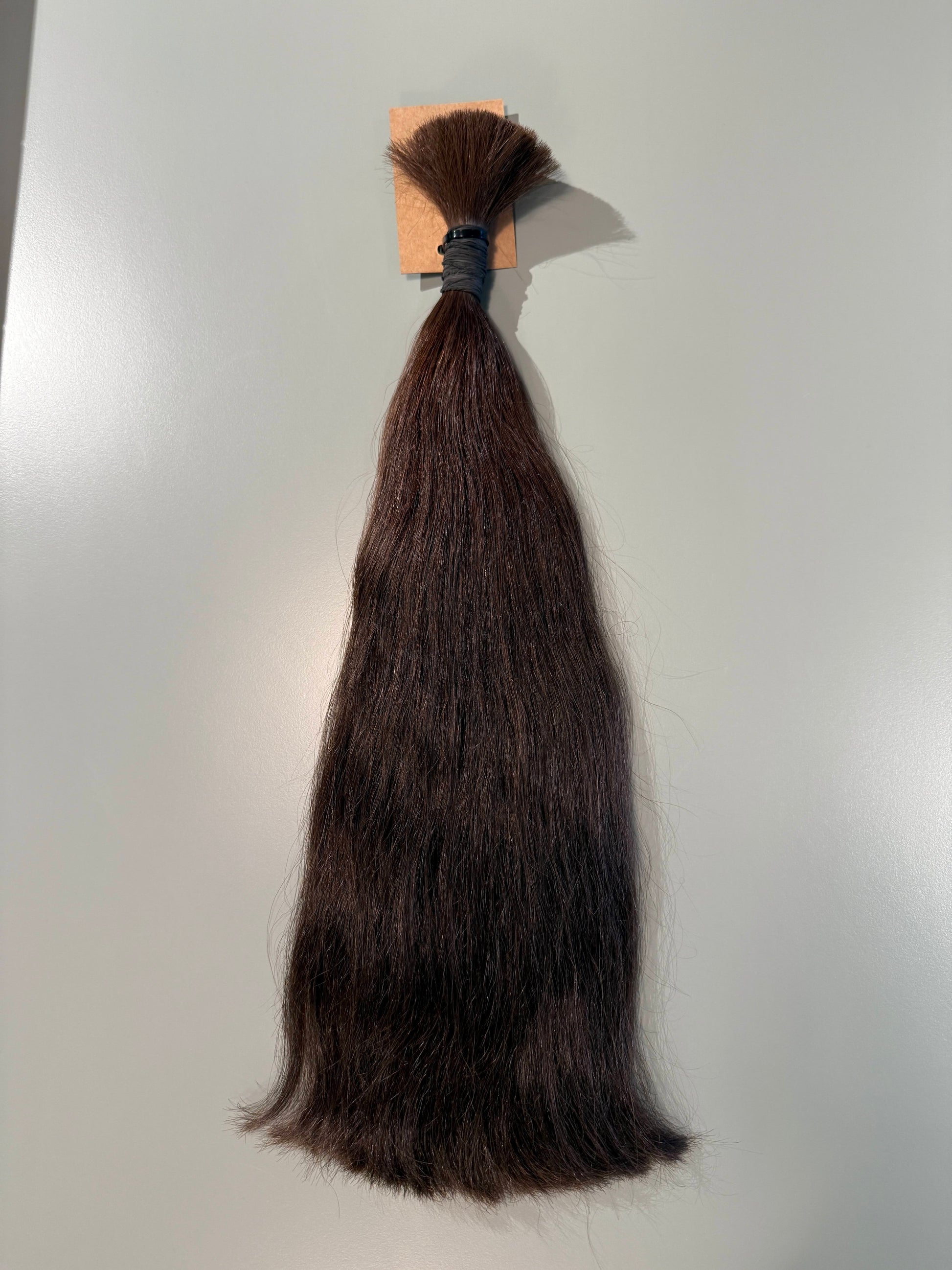 A bundle of straight, dark brown Slavic hair extensions tied with a black elastic band and attached to a small beige cardboard header, shown against a plain light gray background.