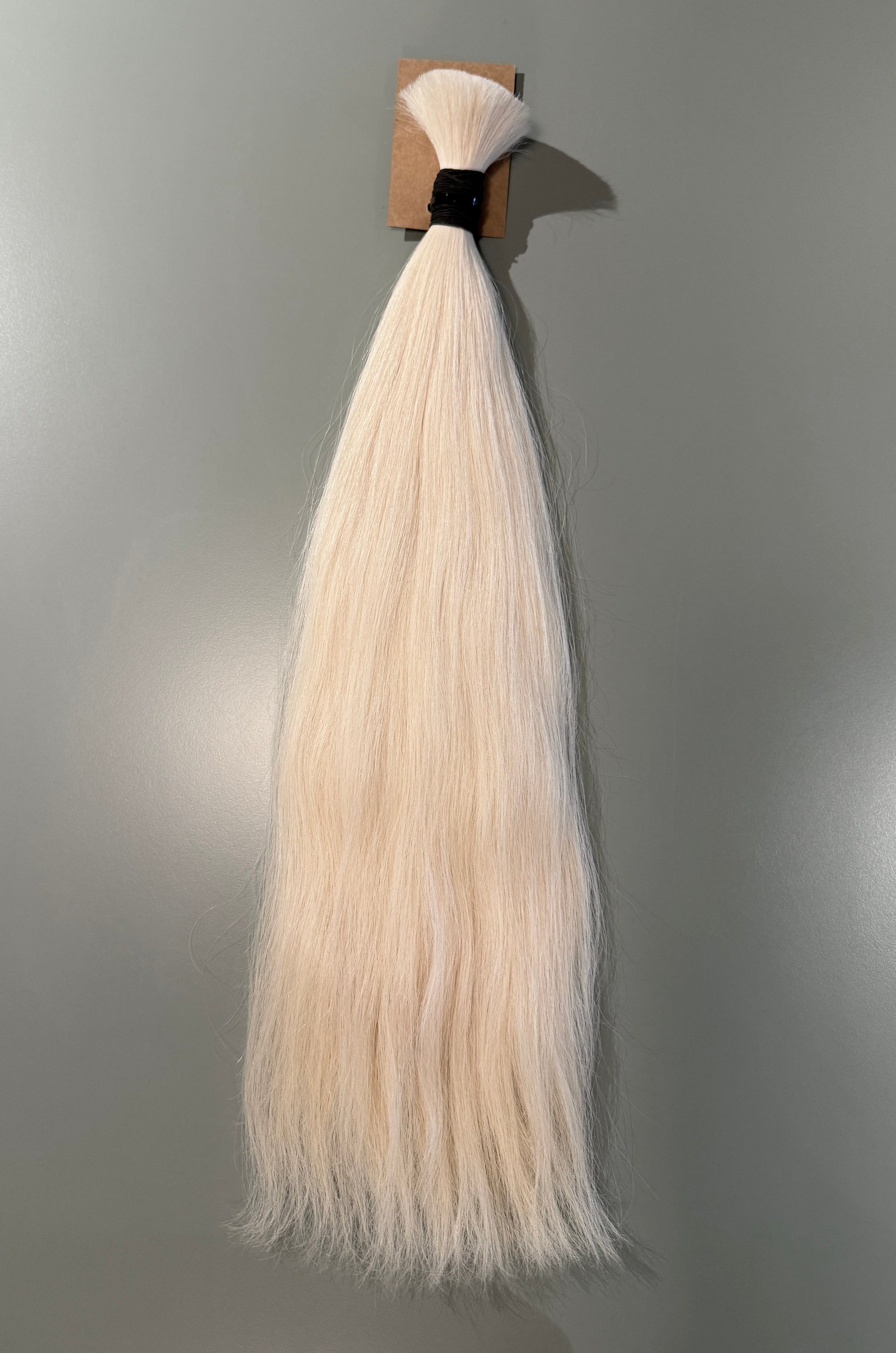 A bundle of long, straight Slavic hair extensions in a very light platinum blonde color, attached to a small cardboard display card with a black band, shown against a plain gray wall.