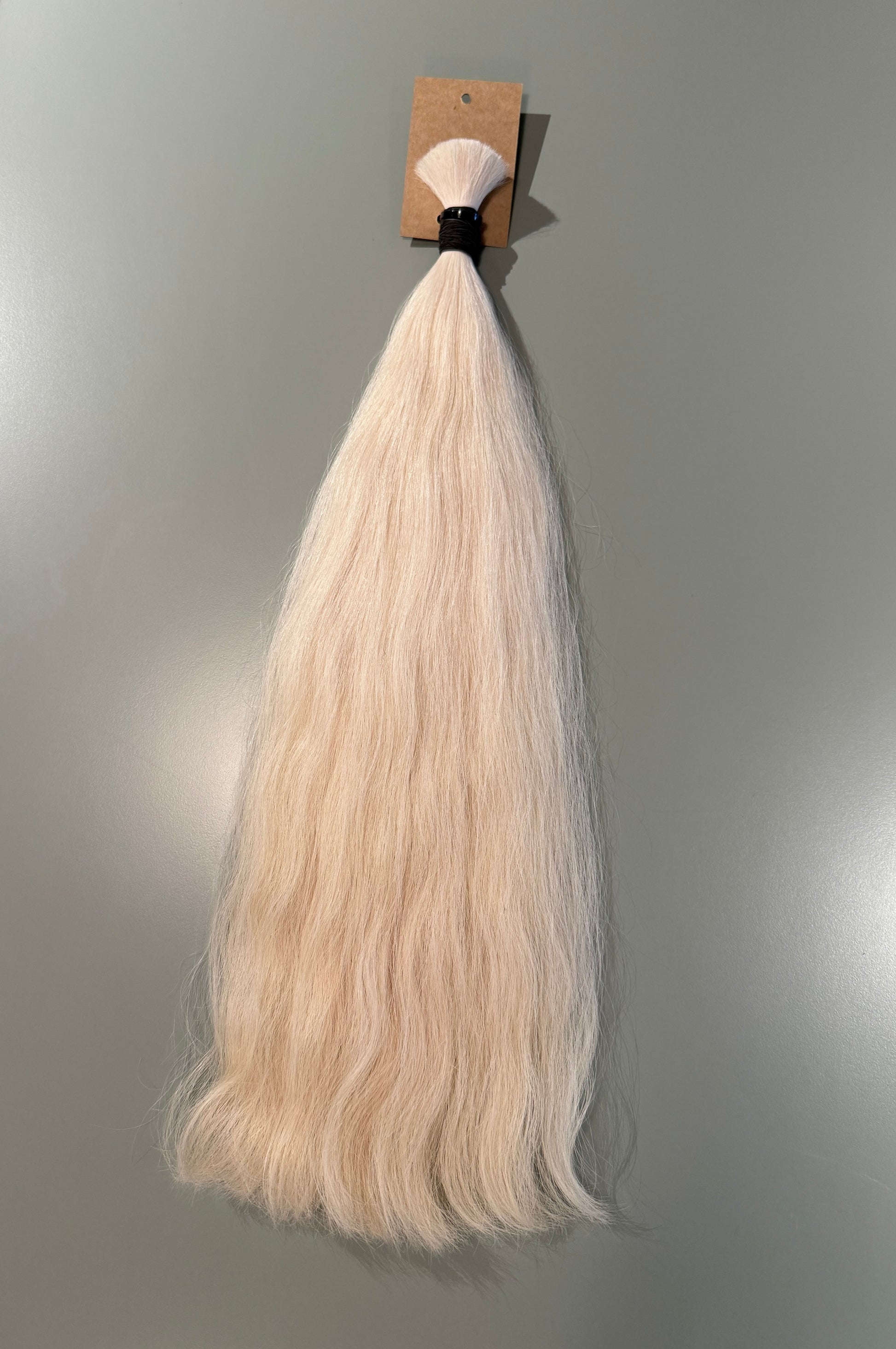 A long, straight slavic hair extension bundle in a very light ash blonde color, attached to a small cardboard display card with a black band, shown against a plain gray wall.