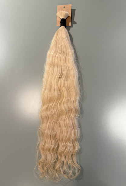 A bundle of long, wavy blonde Slavic hair extensions hanging from a small cardboard display card against a plain gray wall.