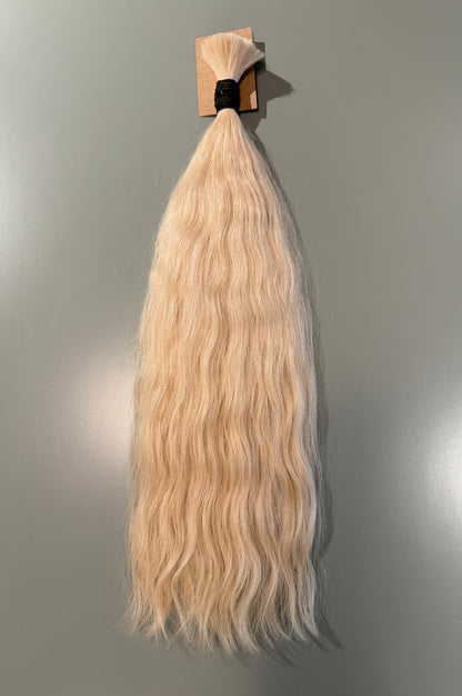 A long, wavy Slavic hair extension piece in a light blonde color, attached to a small cardboard display card with a black elastic band, shown against a plain gray wall.