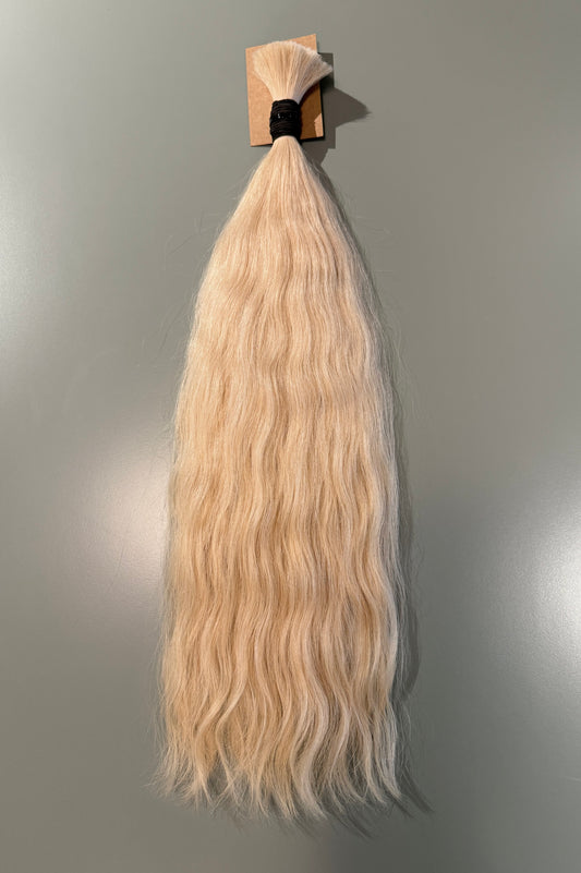 A long, wavy Slavic hair extension piece in a light blonde color, attached to a small cardboard display card with a black elastic band, shown against a plain gray wall.