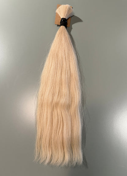 A single bundle of straight, light blonde Slavic hair extensions tied at the top with a black elastic band and small cardboard backing, shown against a plain gray wall.