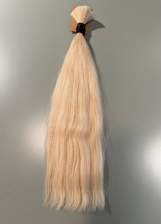 A single bundle of straight, light blonde Slavic hair extensions tied at the top with a black elastic band and small cardboard backing, shown against a plain gray wall.