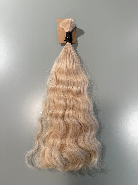 A bundle of long, wavy Slavic hair extensions in a light blonde shade, attached to a small cardboard backing with a black elastic band, shown against a plain gray background