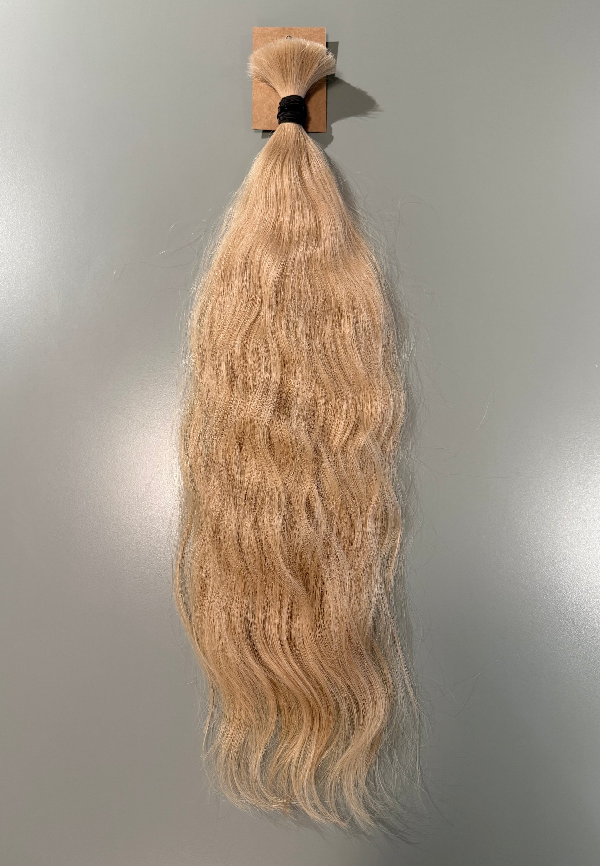 A bundle of long, wavy blonde Slavic hair extensions hanging from a small cardboard holder against a plain light gray wall.