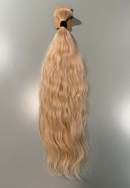 A bundle of long, wavy blonde Slavic hair extensions hanging from a small cardboard holder against a plain light gray wall.