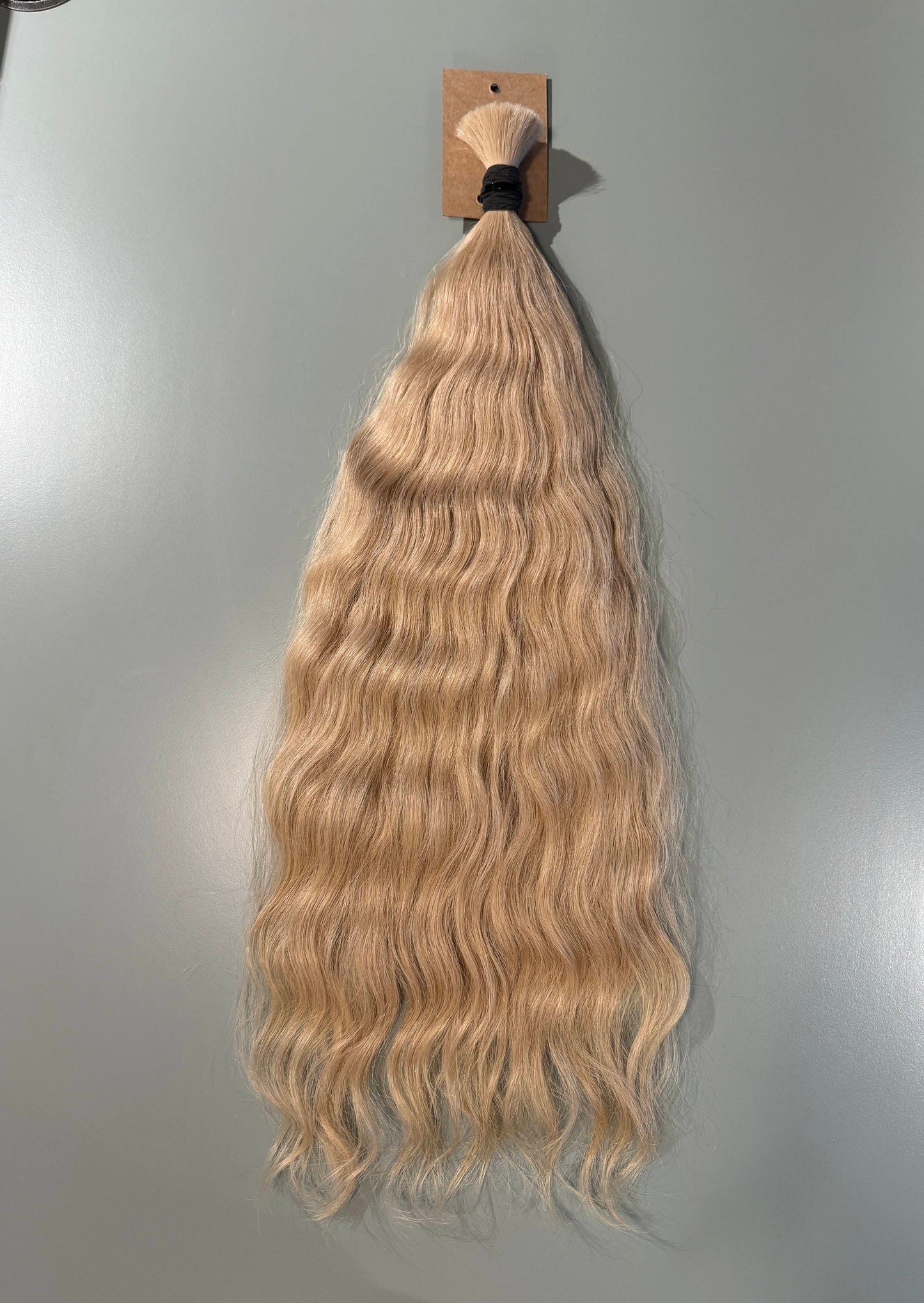 Long wavy blonde Slavic hair extension bundle hanging from a small cardboard display card against a plain light gray wall.