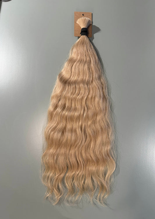 Long wavy blonde Slavic hair extension bundle hanging from a small cardboard display card against a plain light gray wall.