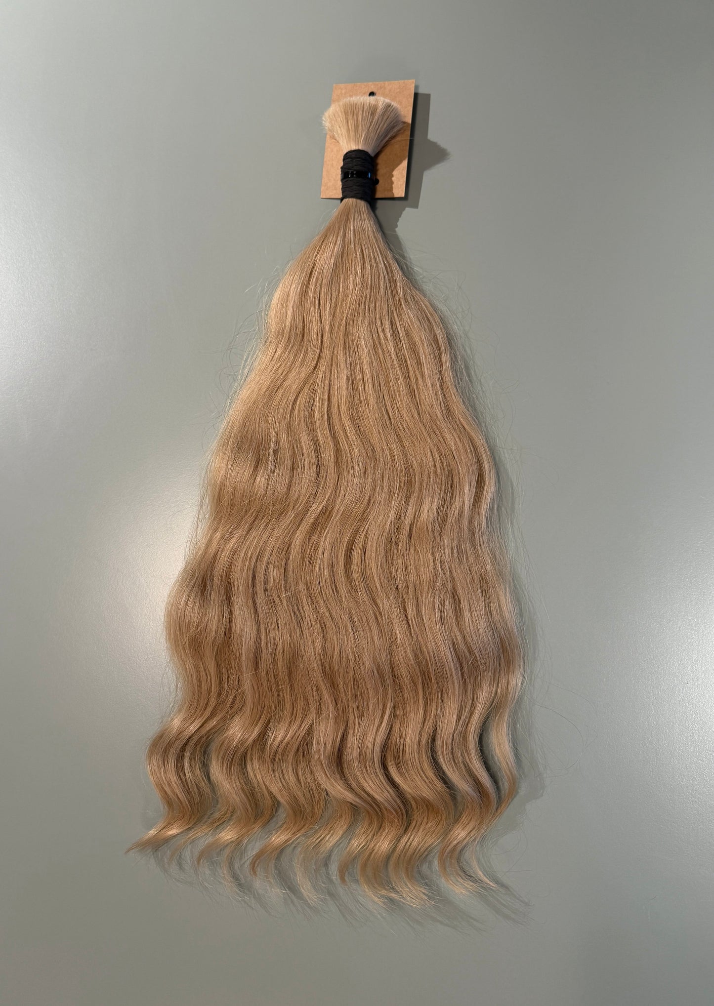 A bundle of long, wavy blonde Slavic hair extensions hanging from a small wooden card against a plain light gray wall.