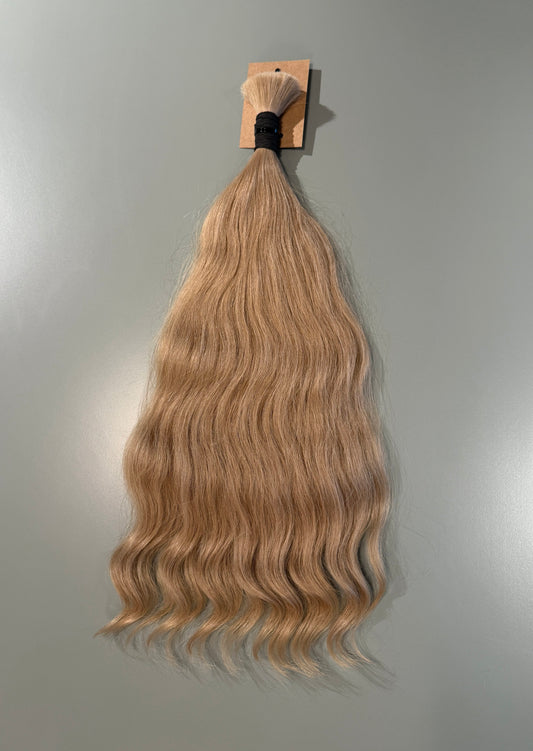A bundle of long, wavy blonde Slavic hair extensions hanging from a small wooden card against a plain light gray wall.