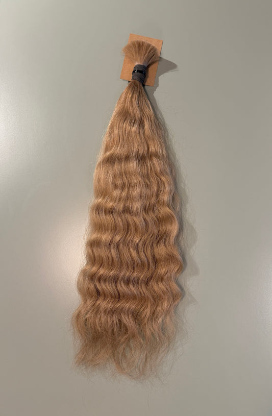 A bundle of wavy, deep-wave Slavic hair extensions in a light golden blonde color, attached to a small cardboard header card with a black elastic band, shown against a plain light gray background.