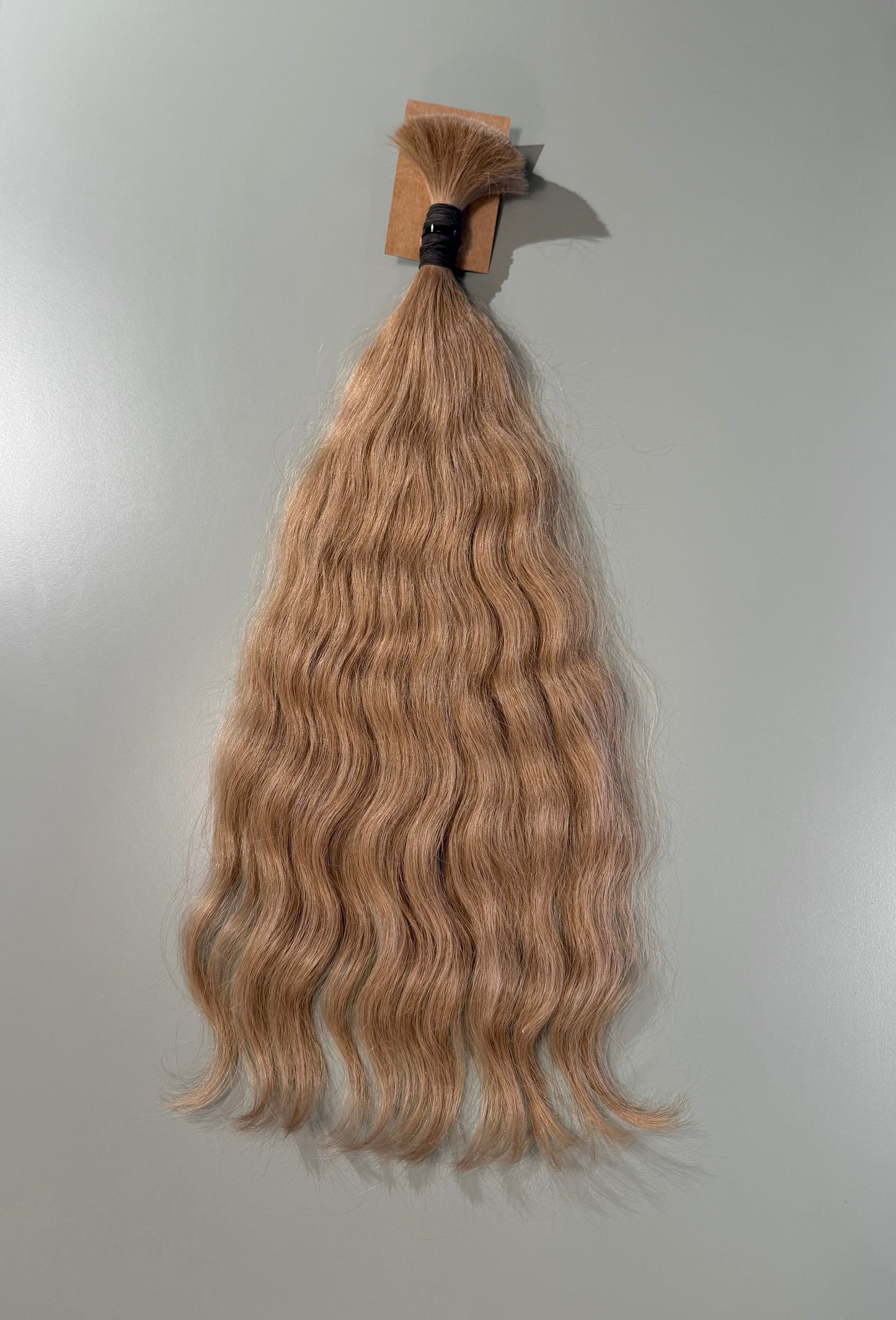 A bundle of wavy, light ash blonde Slavic hair extensions hanging against a plain light gray background.