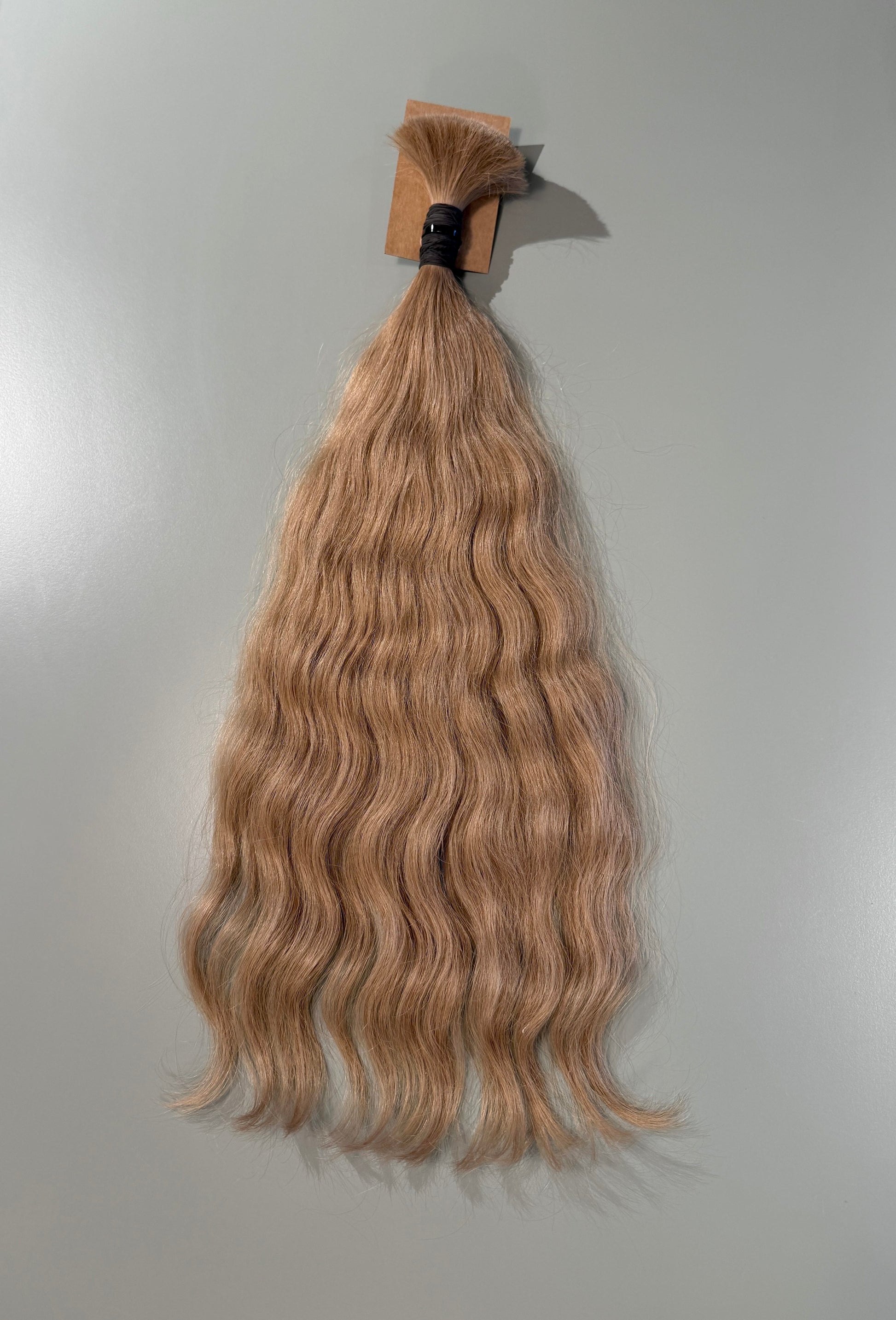 A bundle of wavy, light ash blonde Slavic hair extensions hanging against a plain light gray background.
