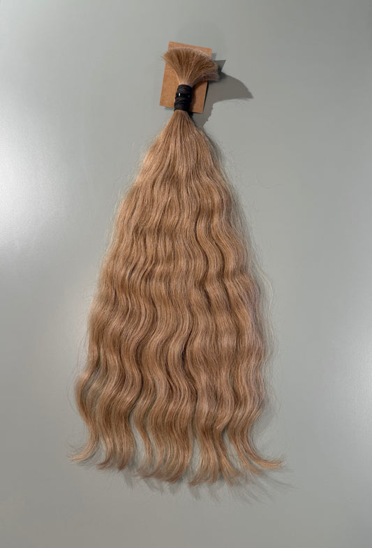 A bundle of wavy, light ash blonde Slavic hair extensions hanging against a plain light gray background.