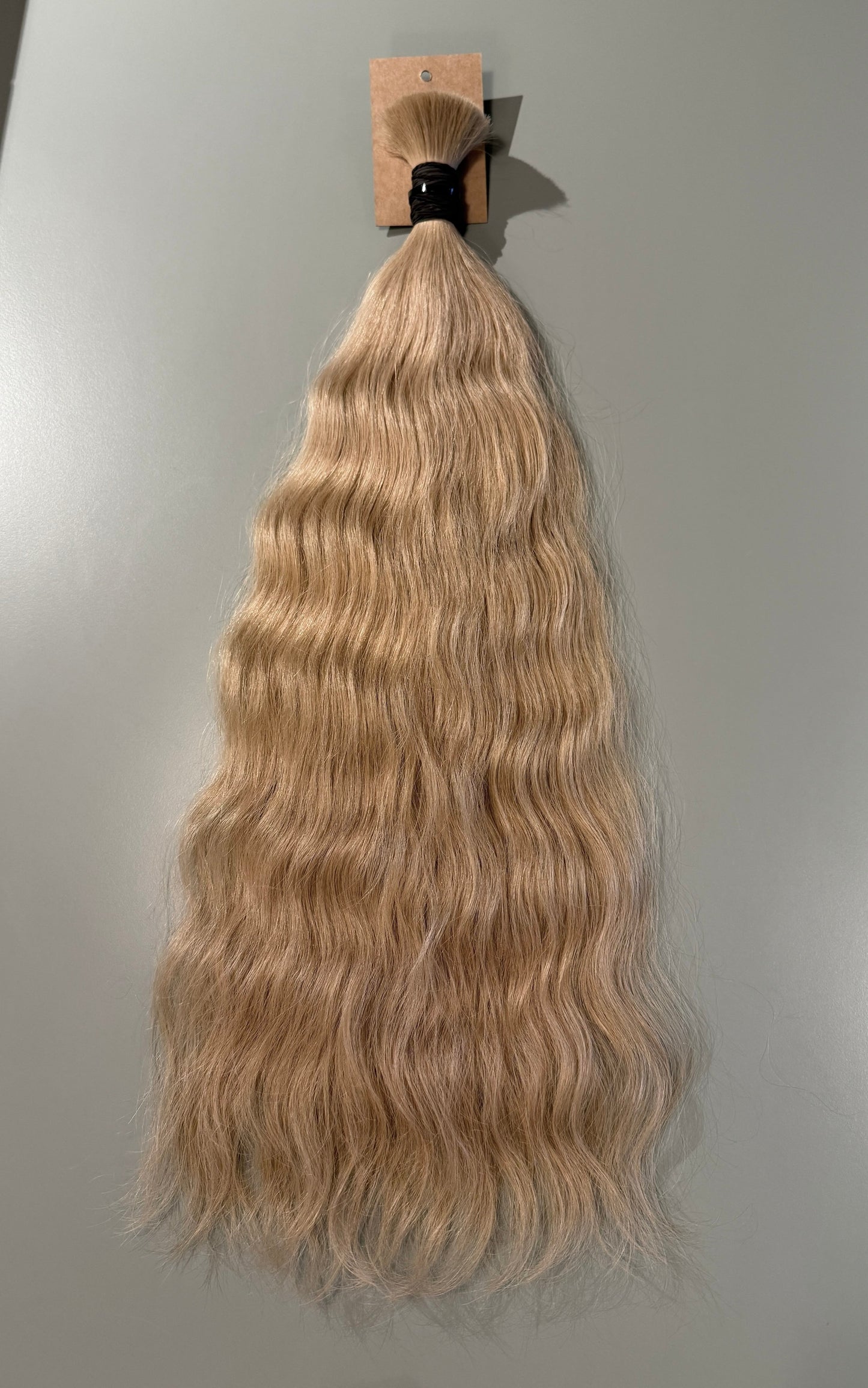 A bundle of long, wavy Slavic hair extensions in a light blonde shade, attached to a small cardboard display card and photographed against a plain light gray background.