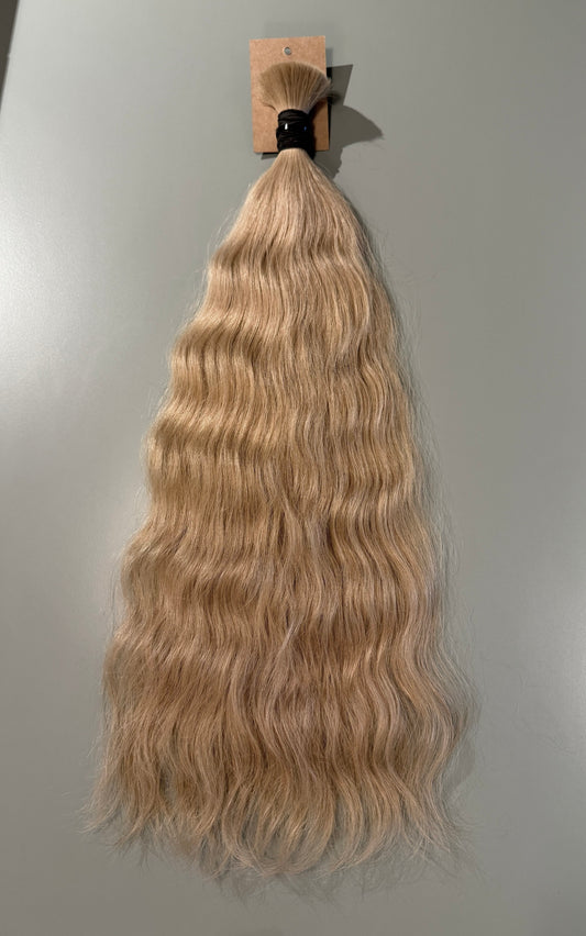 A bundle of long, wavy Slavic hair extensions in a light blonde shade, attached to a small cardboard display card and photographed against a plain light gray background.