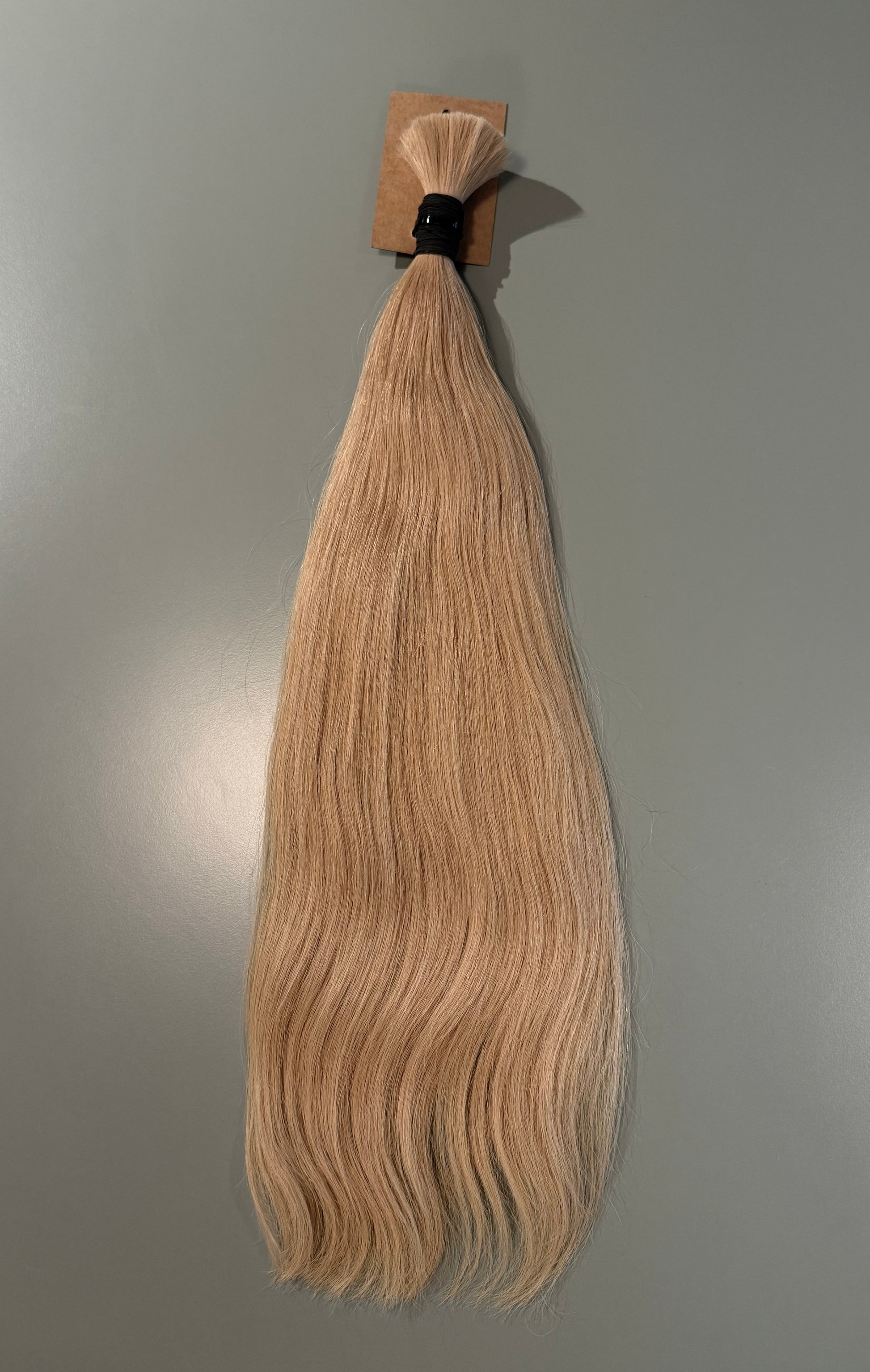 A single bundle of straight, wavy Slavic blonde hair extensions attached to a small cardboard header with a black elastic band, shown against a plain gray background.