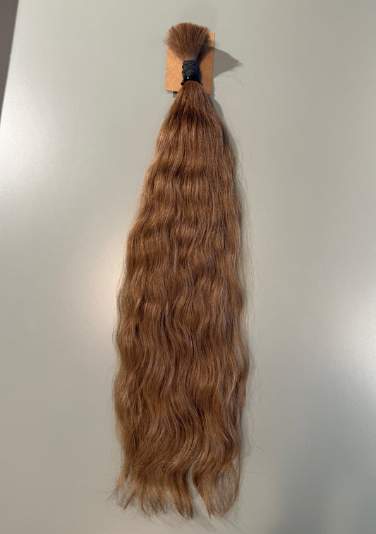 A bundle of long, wavy Slavic hair extensions in a light ash brown shade, attached to a cardboard header with a black elastic band, shown against a plain light gray background.