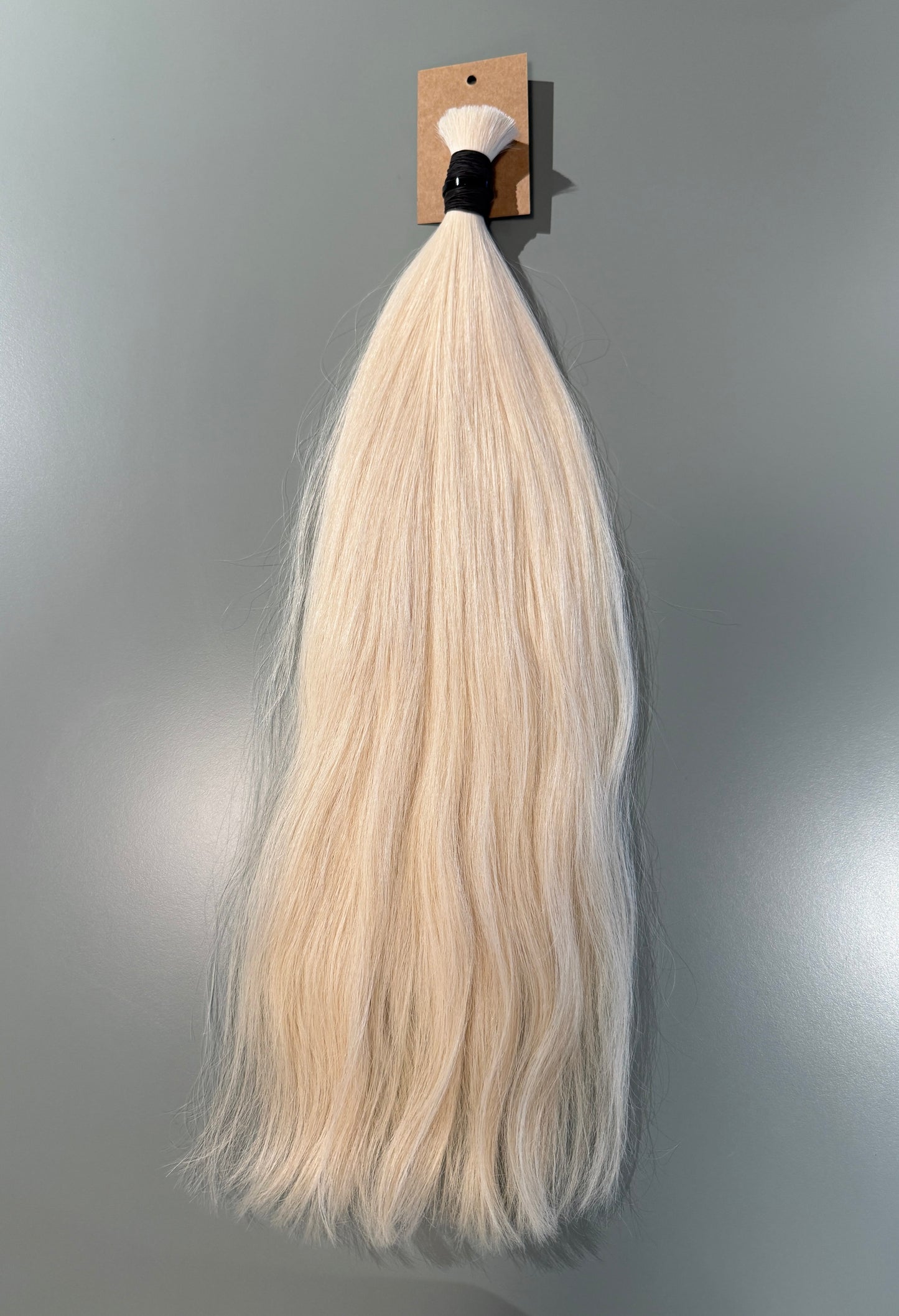 A long, straight slavic hair extension bundle in a very light platinum blonde color, attached to a small cardboard display card with a black band, shown against a plain gray wall.